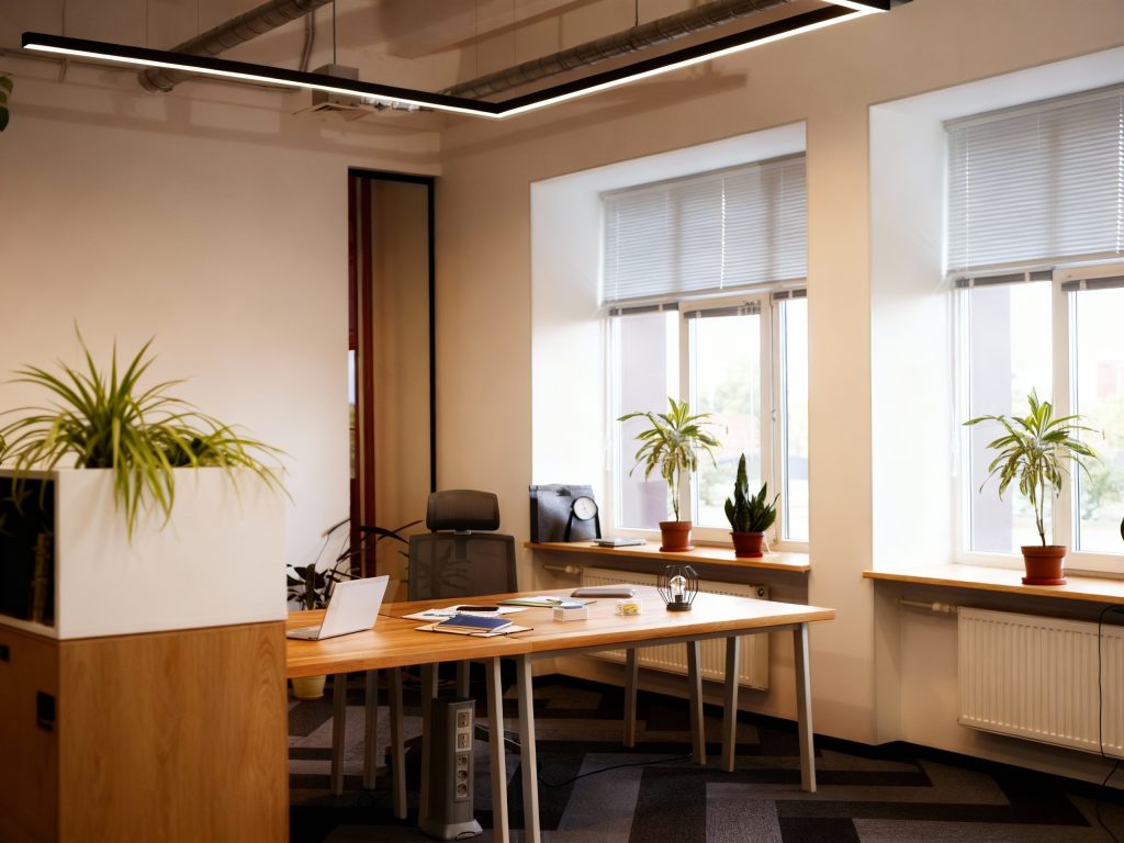 Nowoczesne, jasne biuro z drewnianymi biurkami i roślinami ustawionymi na parapetach; w oknach zamontowane są rolety wewnętrzne w jasnym kolorze, które rozpraszają światło dzienne i zapewniają komfort pracy przy stanowisku z laptopem i dokumentami.