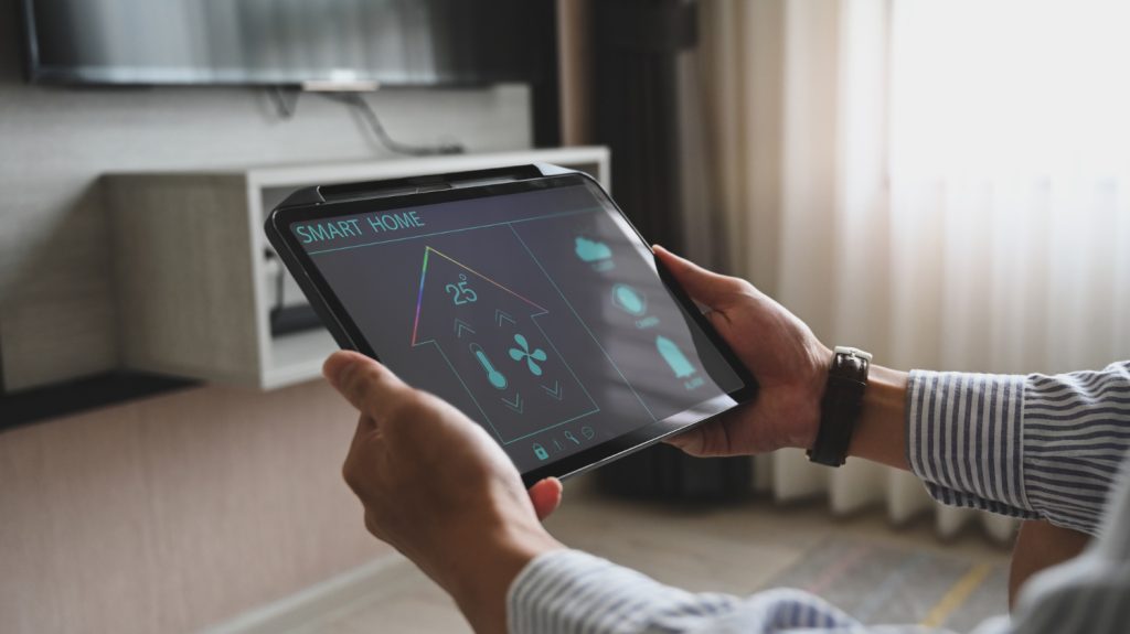 Osoba trzymająca tablet i obsługująca system smart home, widoczny ekran wyświetlający temperaturę 25°C oraz ikony sterujące urządzeniami - wentylatorem, alarmem czy roletami zewnętrznymi.