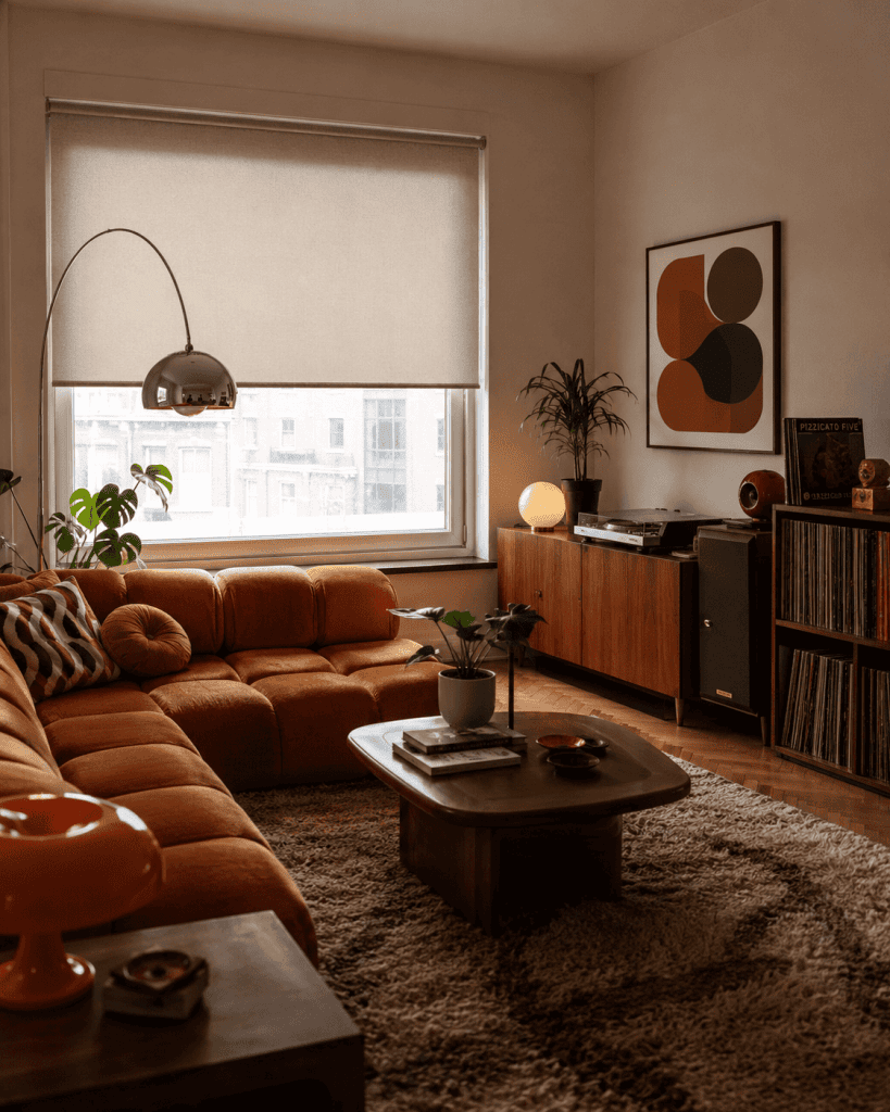 Salon vintage z pomarańczową sofą, drewnianymi meblami, kolekcją płyt winylowych i jasną roletą wewnętrzną.