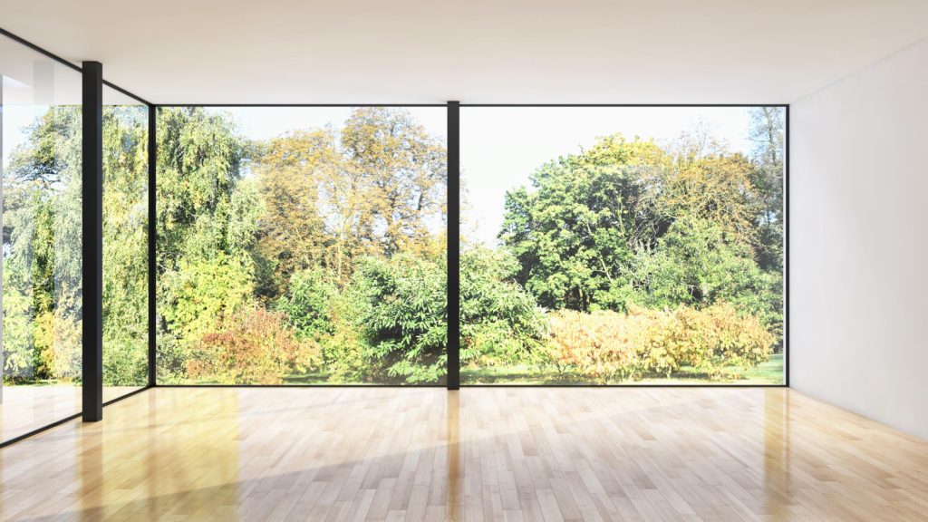 Przestronne wnętrze z jasną podłogą i dużymi oknami, przez które widać ogród z drzewami i krzewami.