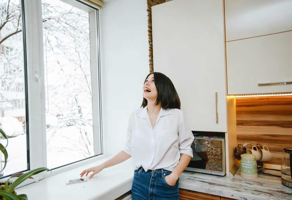 Kobieta stoi w kuchni przy oknie z roletą, patrząc na nią. W tle widoczne są szafki kuchenne i blat z naczyniami.